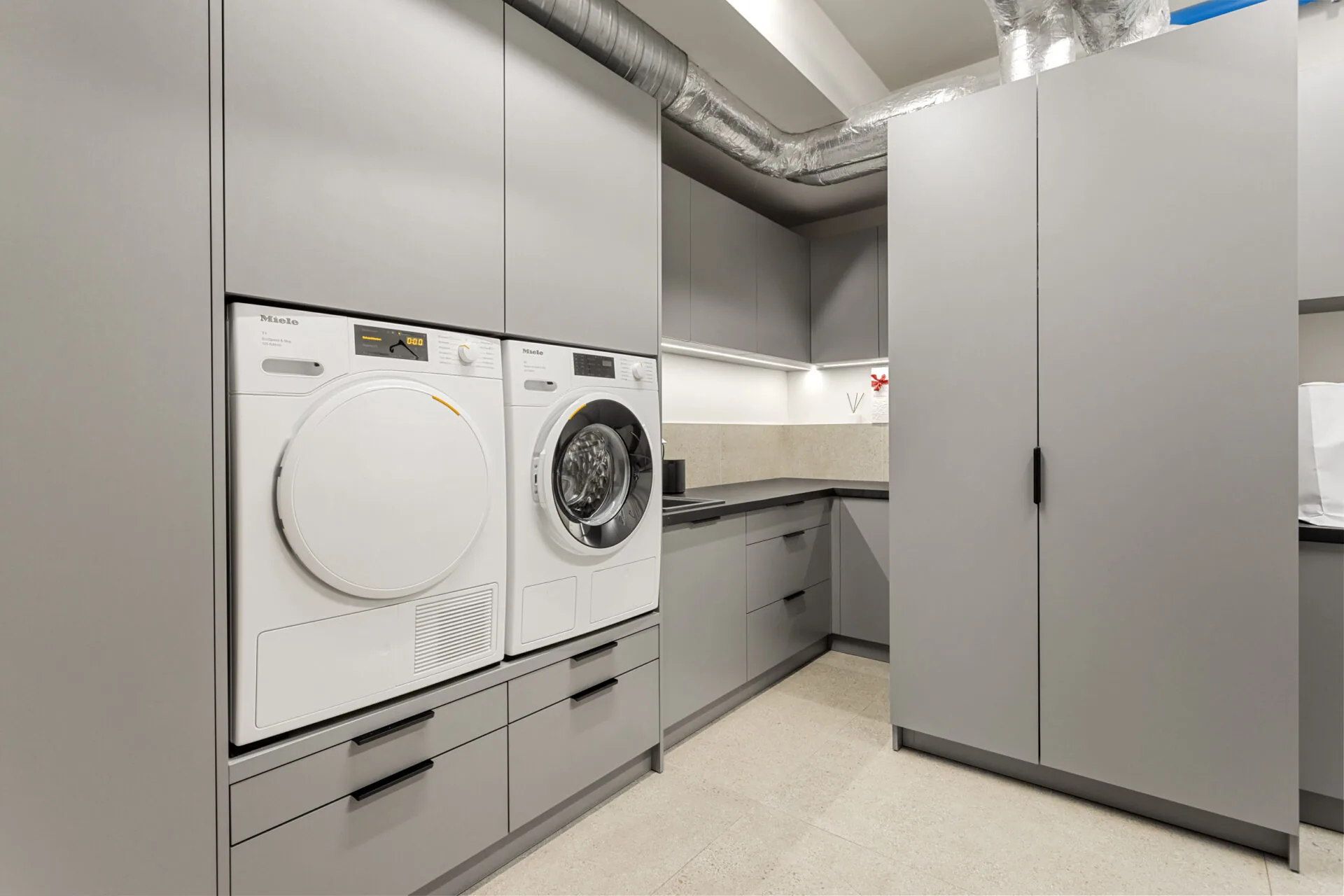 Hauswirtschaftsraum mit grauen Schrankfronten und Waschmaschine mit Trockner auf einem Sockel mit Schubladen